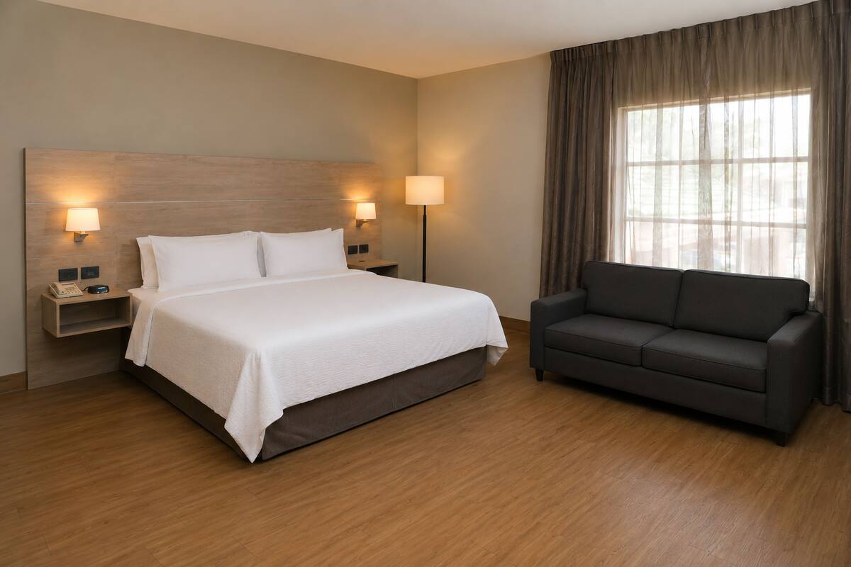A hotel room with a bed, bedside lamps, a nightstand, a sofa, and a large window with curtains.