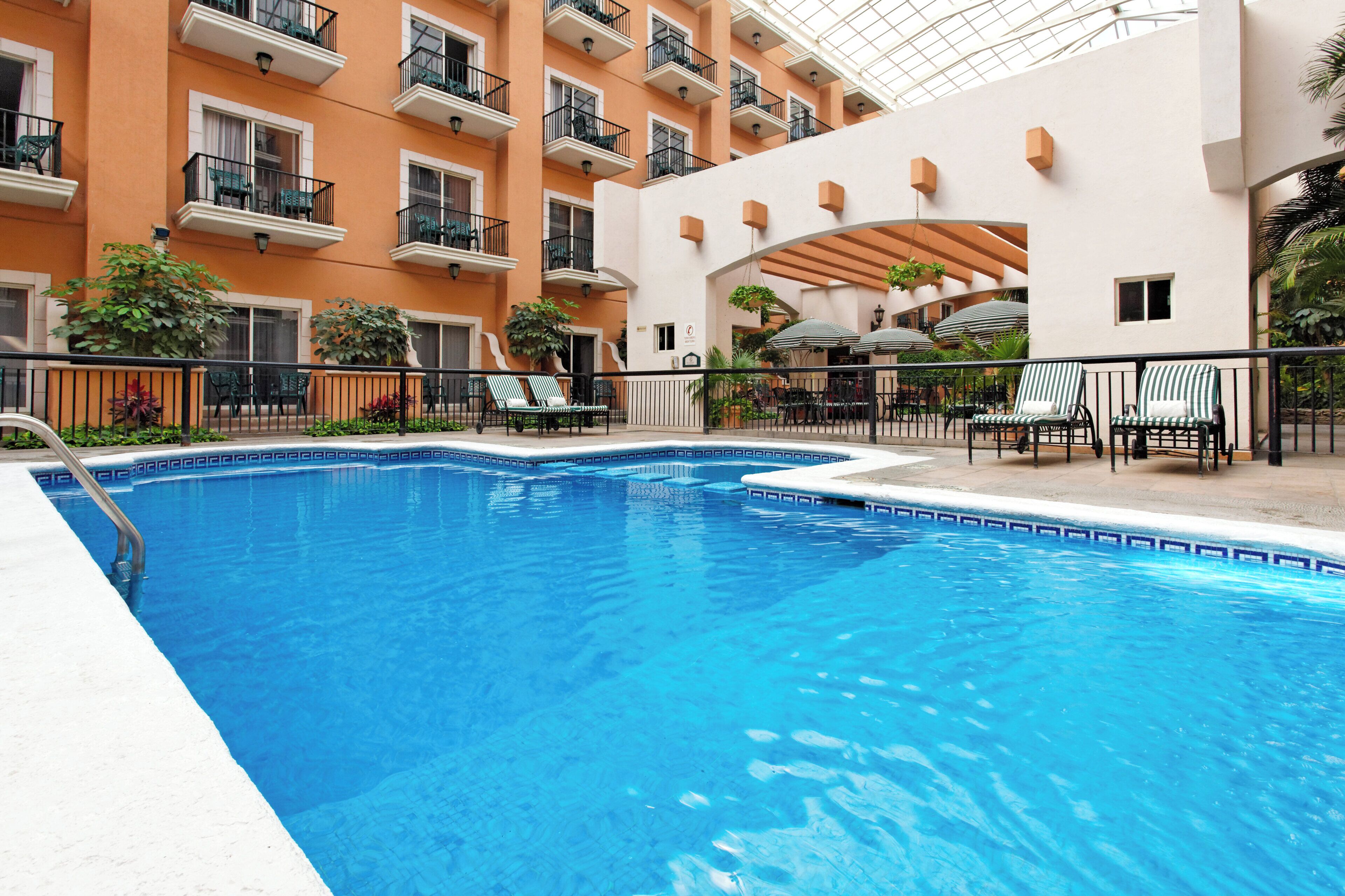 Una piscina con sillas de descanso y sombrillas, rodeada por un edificio con balcones y un área de patio techada.