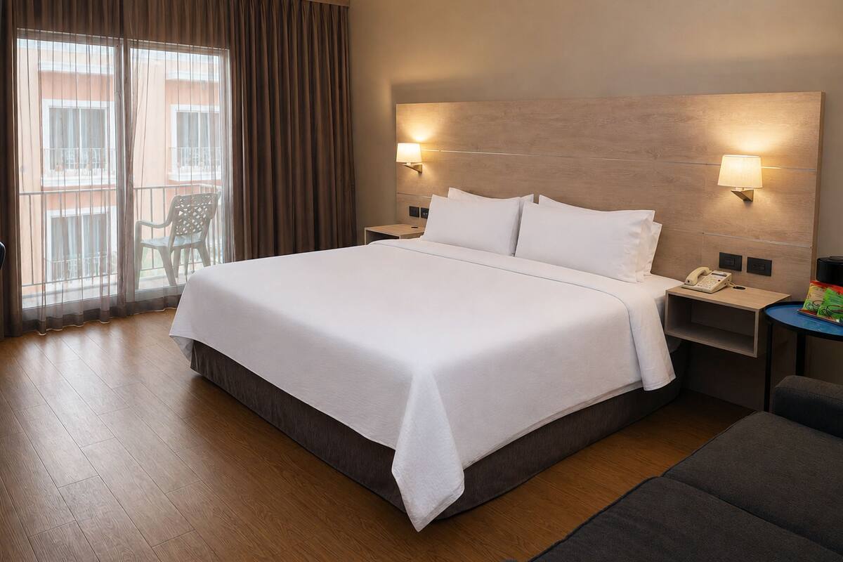 A hotel room with a large bed, two bedside lamps, a telephone, and a view of the outside through a window with curtains.