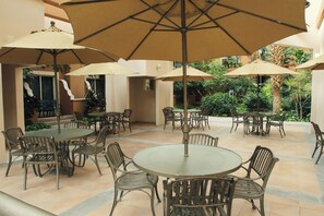 Un espacio al aire libre con mesas y sillas redondas, rodeado de sombrillas color beige.