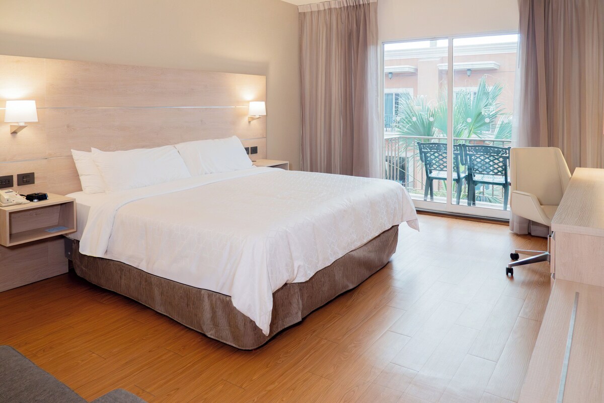 A hotel room with a large bed, bedside tables, a chair, and a balcony with a view.