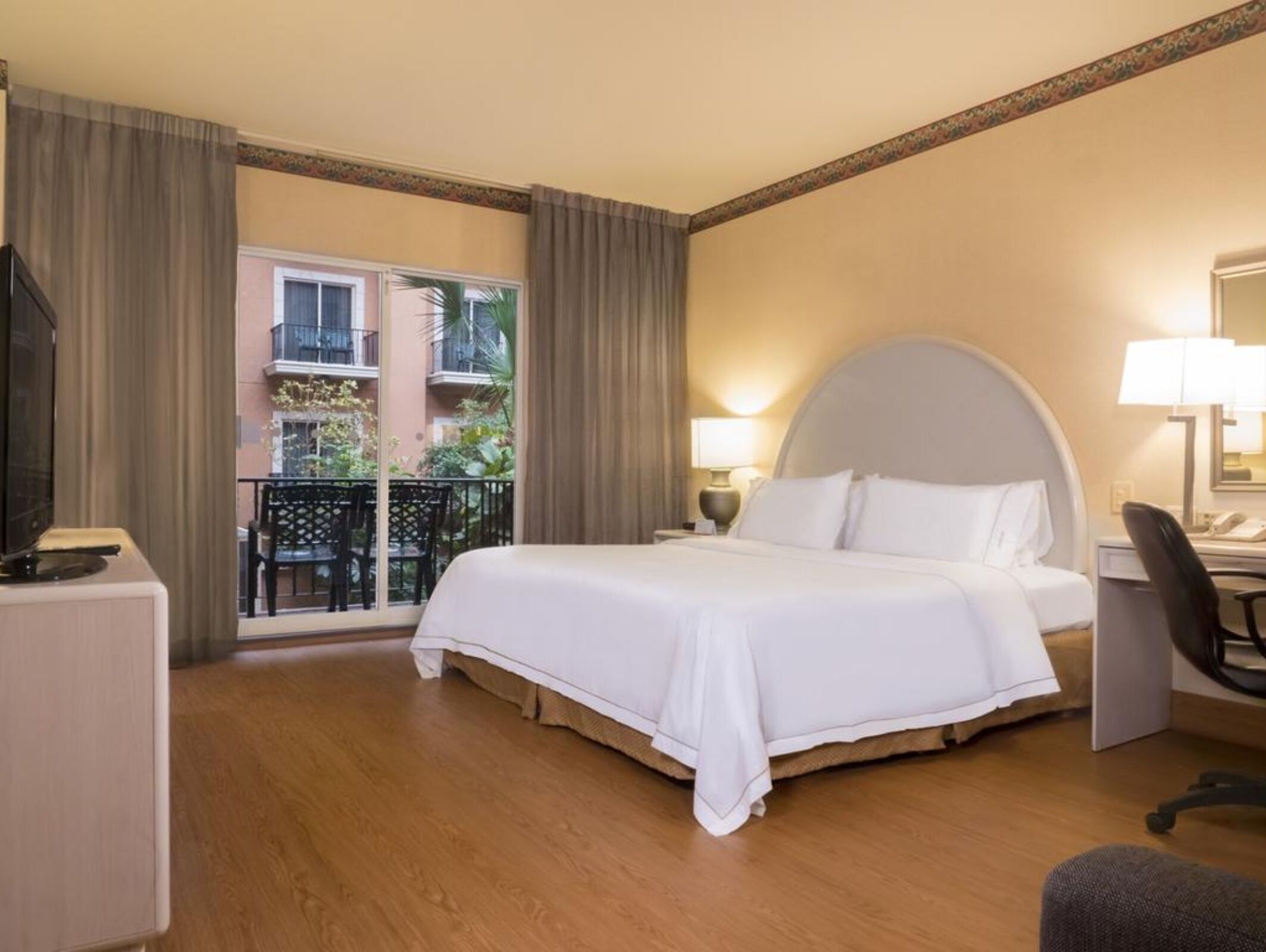 A hotel room with a large bed, a desk, a chair, and a balcony with a view of buildings and greenery.