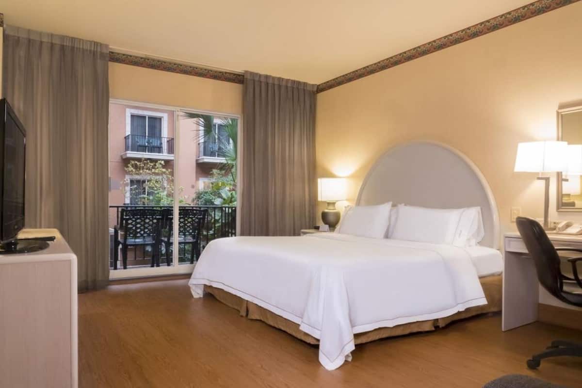 Une chambre d’hôtel avec un grand lit, un bureau, une chaise et un balcon offrant une vue sur les bâtiments et la verdure.