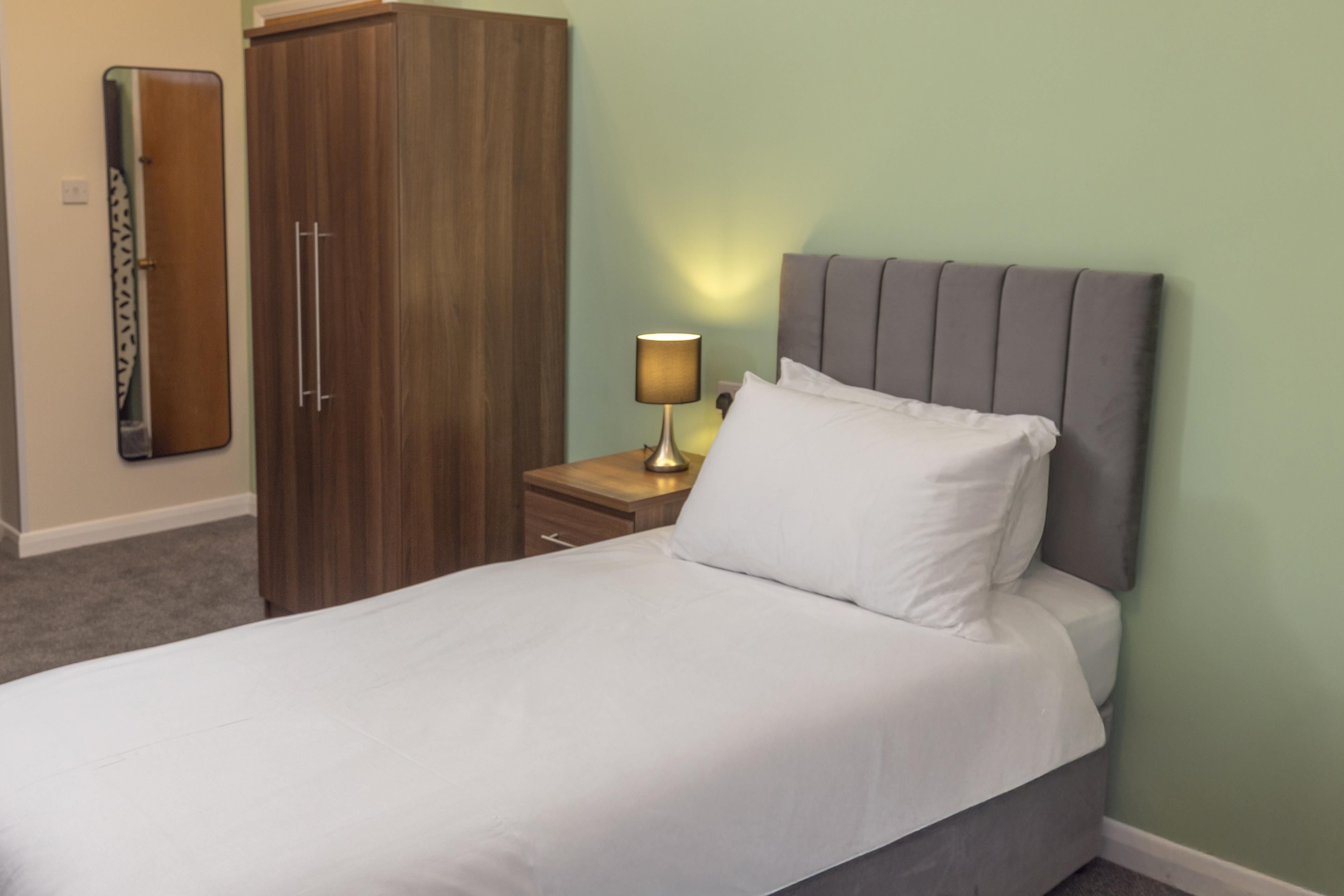 A neatly made bed with white linens, a bedside lamp, and a wooden wardrobe in a room with green walls.