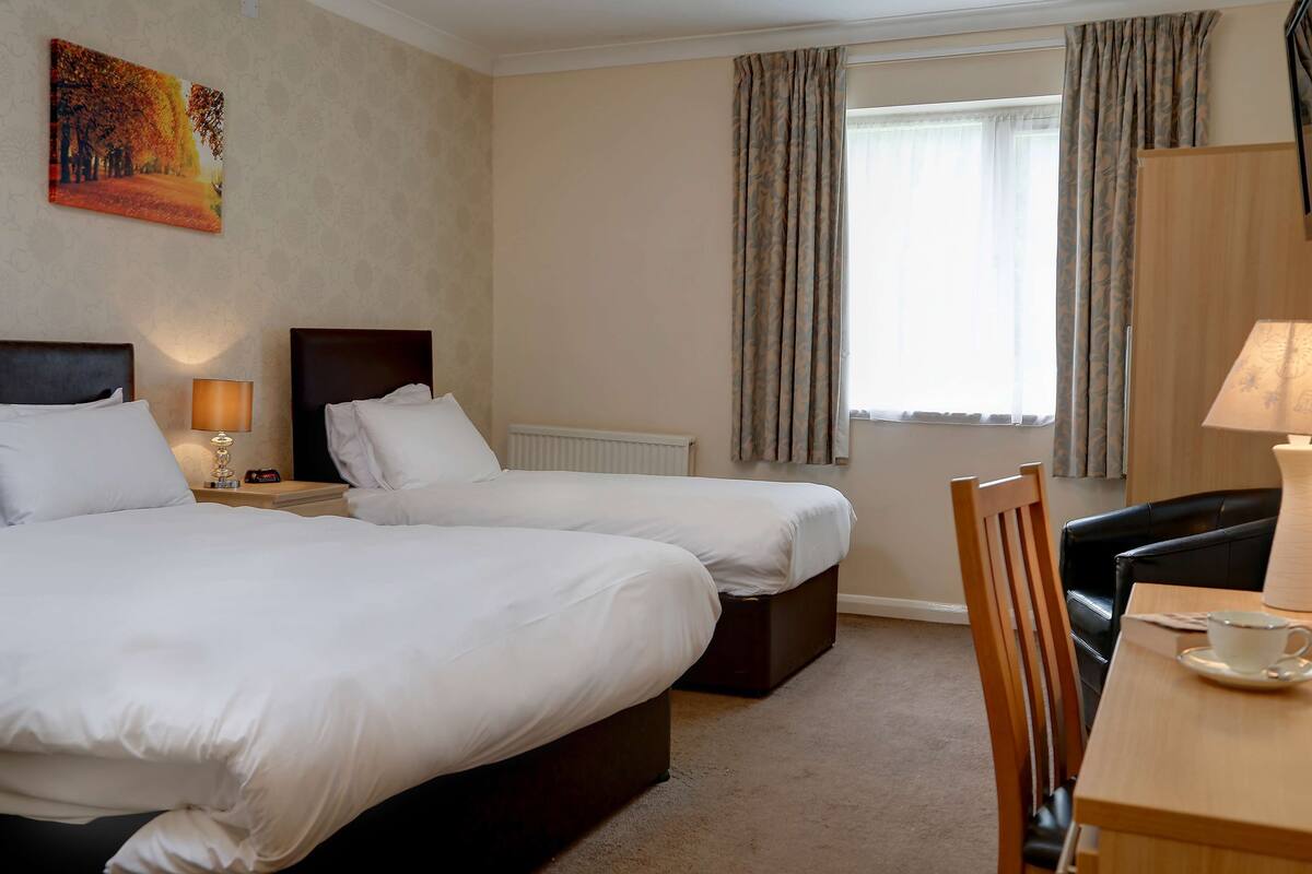 A hotel room with two beds, a wooden chair, a table with a cup, and a window with curtains.