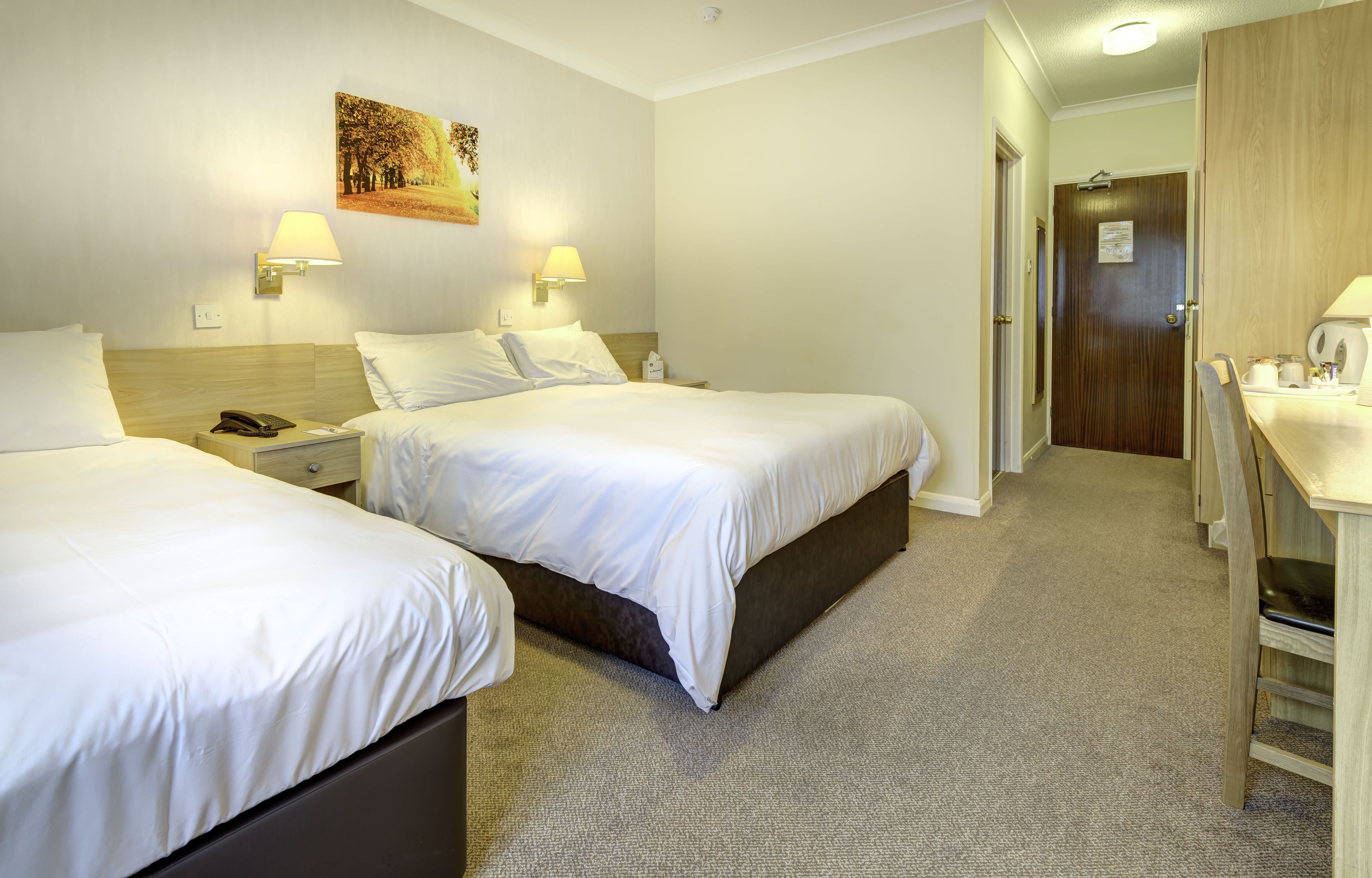 A hotel room with two beds, a desk, and a chair. There are two wall-mounted lamps and a painting on the wall.