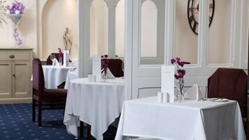 A dining area with tables set for two, white tablecloths, and purple flowers in vases.
