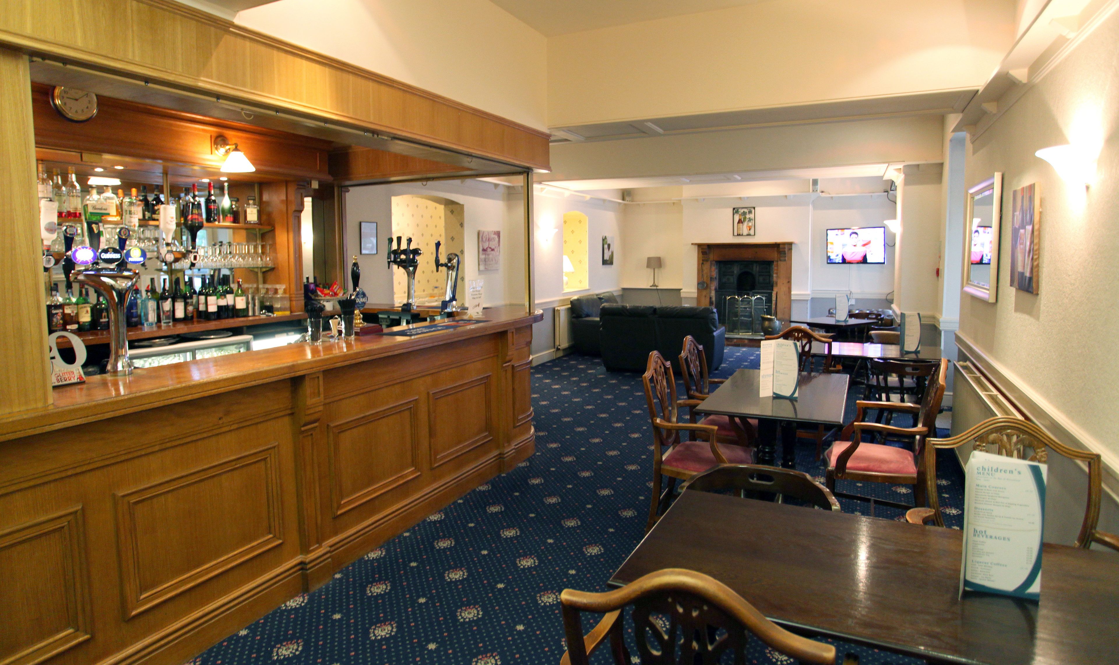 A bar area with a wooden counter, multiple taps, and a variety of bottles. There is a seating area with tables and chairs, a television, and a fireplace.