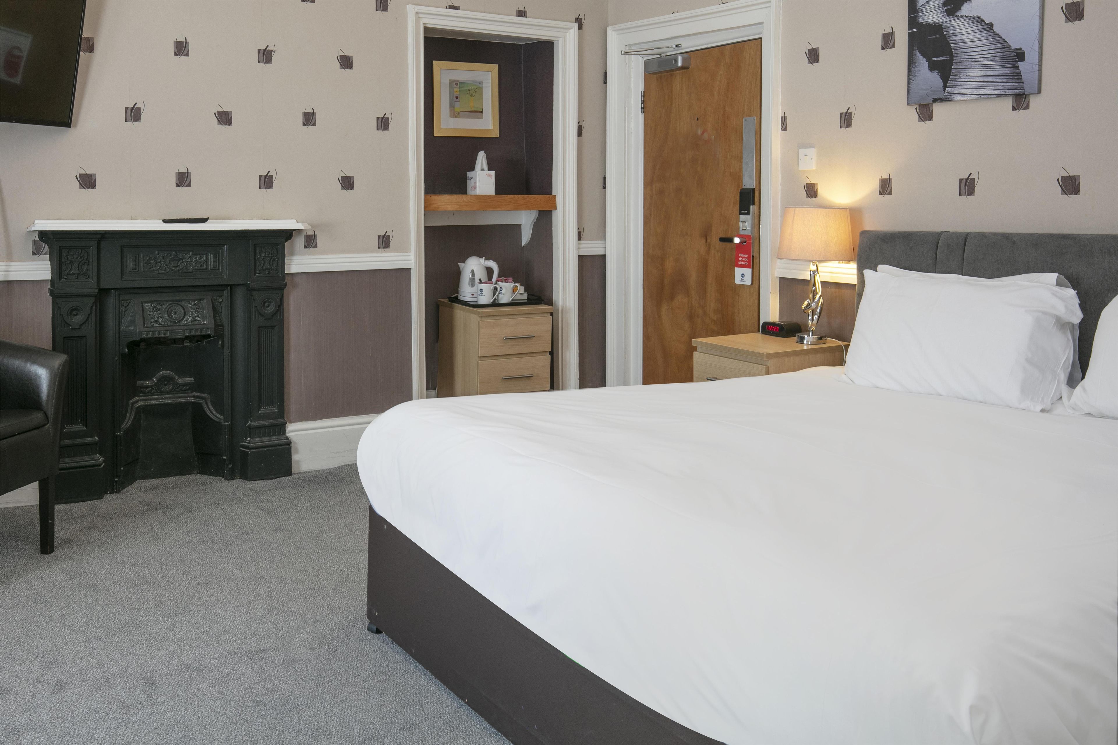 A hotel room with a large bed, a desk with a kettle, a wooden cabinet, and a nightstand with a lamp.