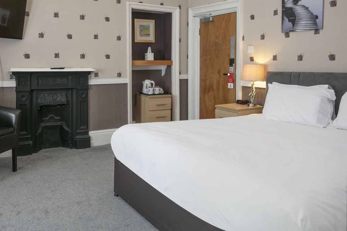 A hotel room with a large bed, a desk with a kettle, a wooden cabinet, and a nightstand with a lamp.