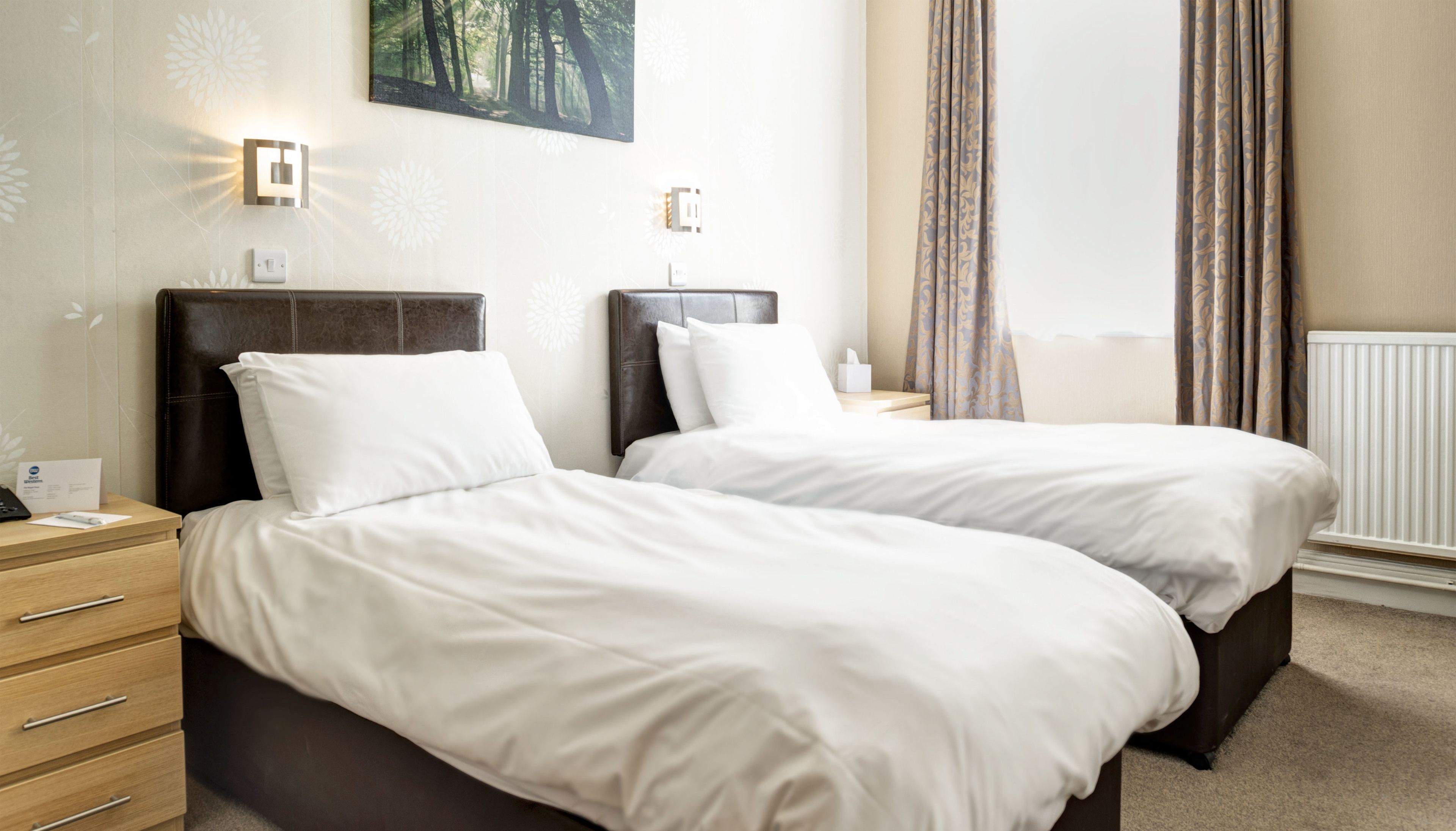 A hotel room with two beds, a nightstand with a lamp, a painting on the wall, and a window with curtains.