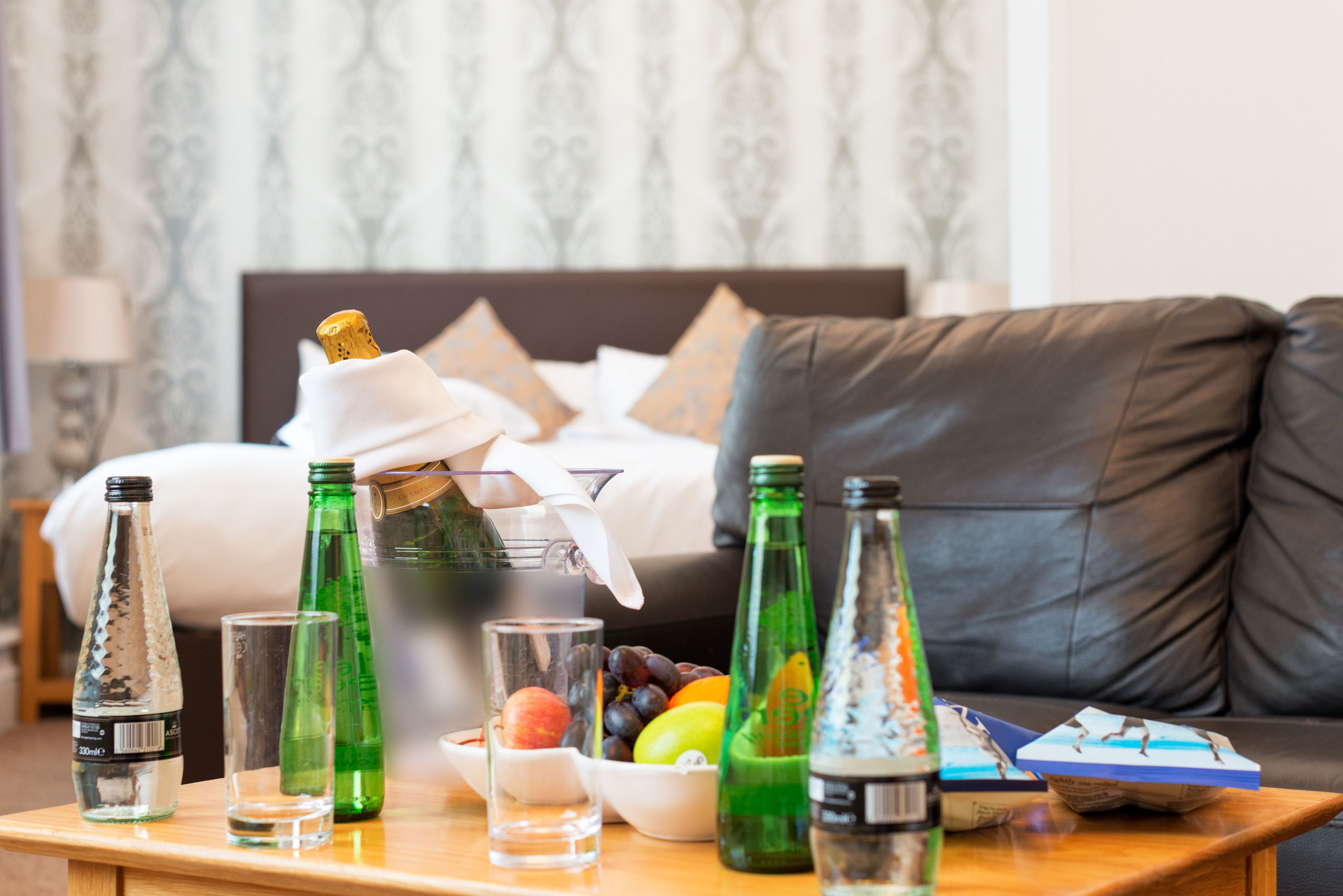 Une chambre d’hôtel avec un lit, une table en bois sur laquelle se trouvent diverses bouteilles et des verres, et un saladier rempli de fruits.