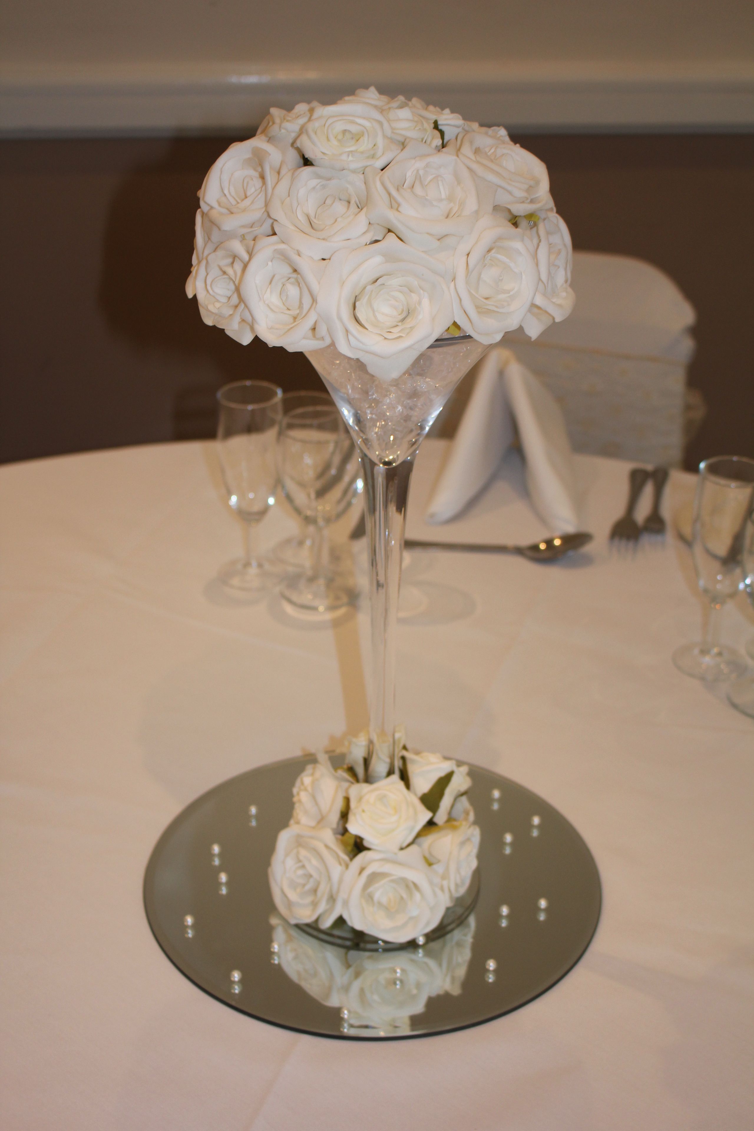 Une table avec un plateau en verre transparent, un grand vase en verre contenant des roses blanches et une serviette blanche pliée.