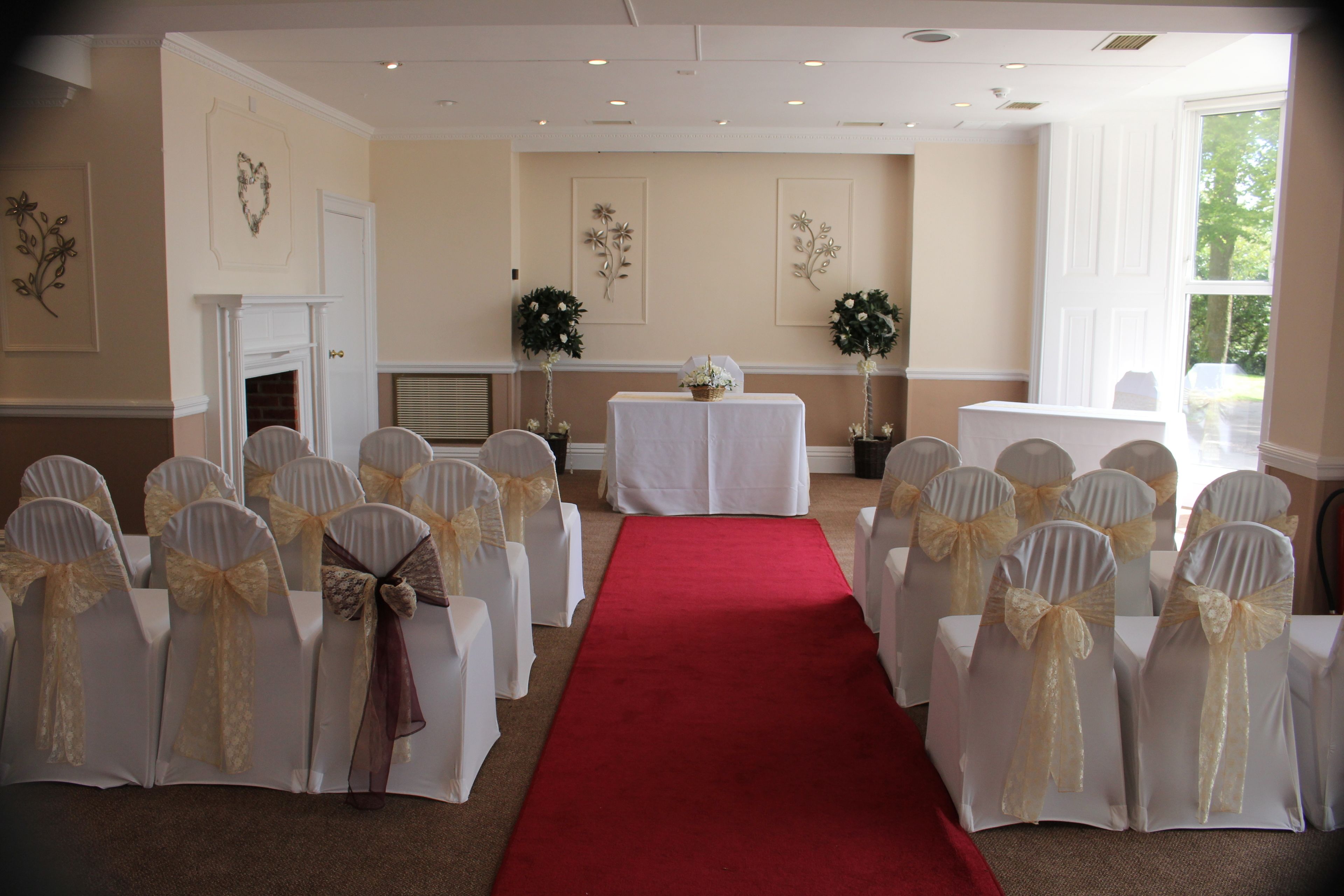 A room with a red carpet aisle, white chairs with gold sashes, and a white table with a floral arrangement.
