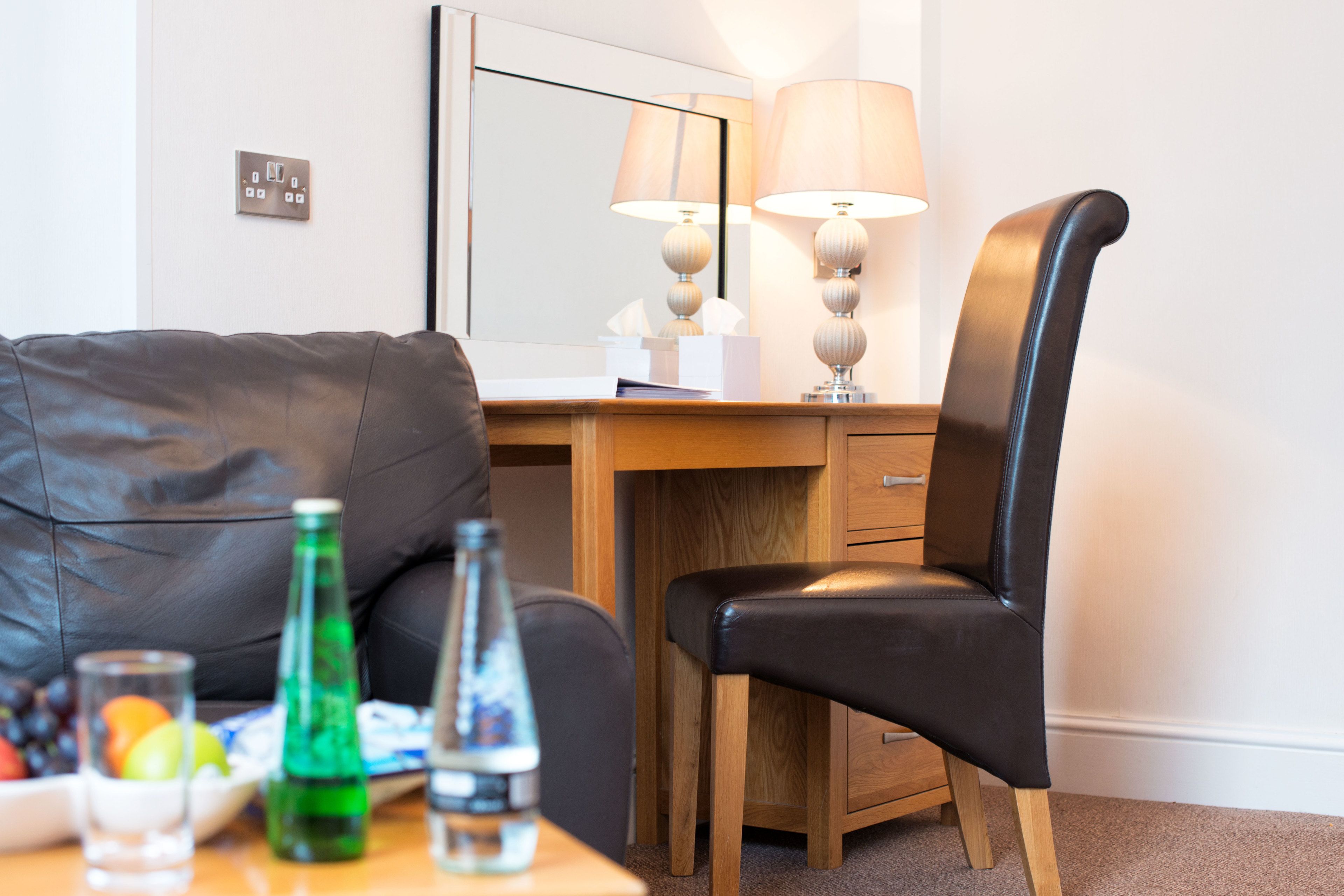 A hotel room with a leather sofa, wooden desk, and a chair. There are bottles and glasses on the table.