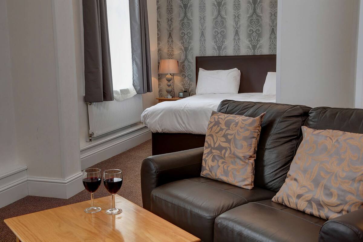 A hotel room with a bed, a leather sofa, and two glasses of red wine on a wooden table.