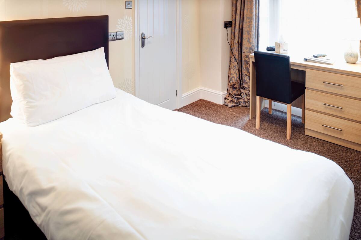 A neatly made bed with white linens, a wooden headboard, a desk with a chair, and a window with curtains.