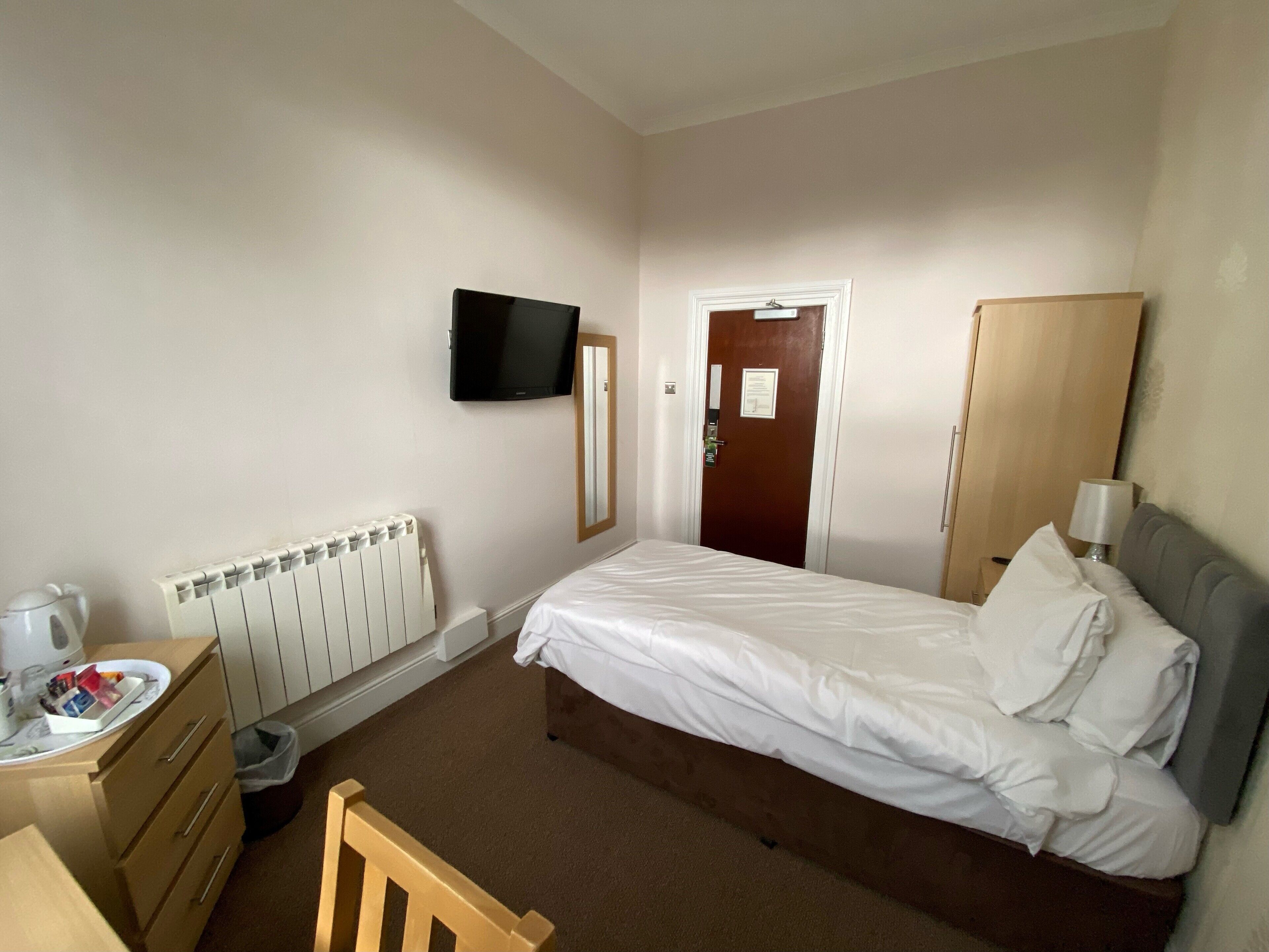 Une chambre d’hôtel avec un lit, un téléviseur fixé au mur, une chaise en bois, une commode et un radiateur.