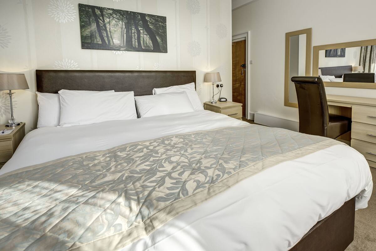 A neatly made bed with a patterned bedspread, a bedside table with a lamp, a nightstand with a phone, a desk with a chair, and a mirror on the wall.