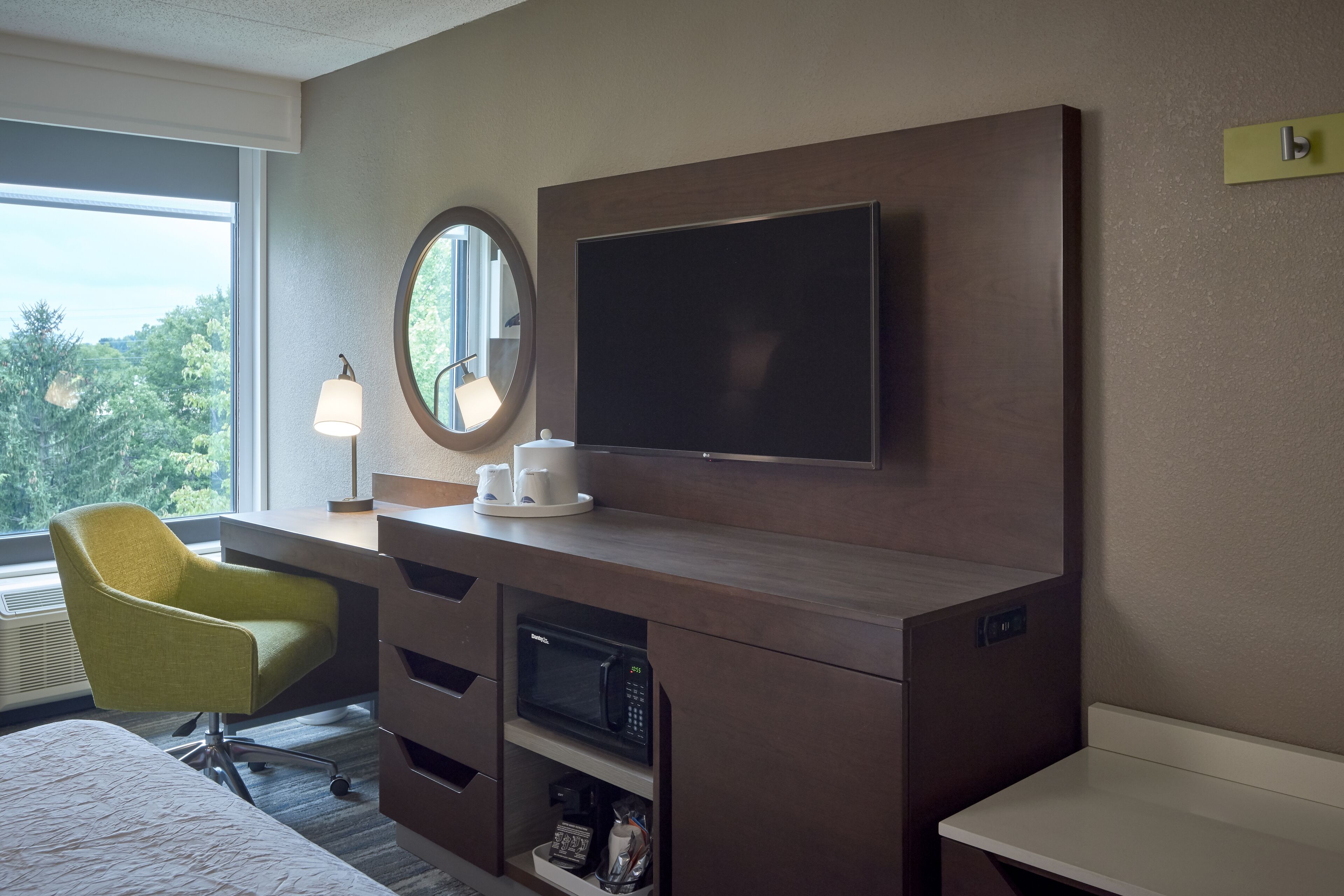 A hotel room with a bed, a desk with a flat-screen TV, a microwave, a chair, and a window with a view of trees.