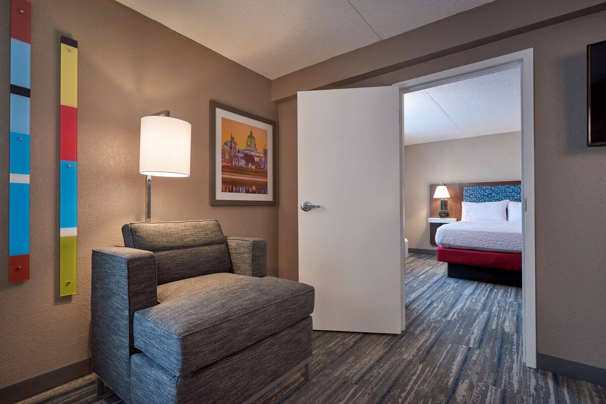 A hotel room with a gray sofa, a white door leading to a bedroom with a bed and a nightstand.