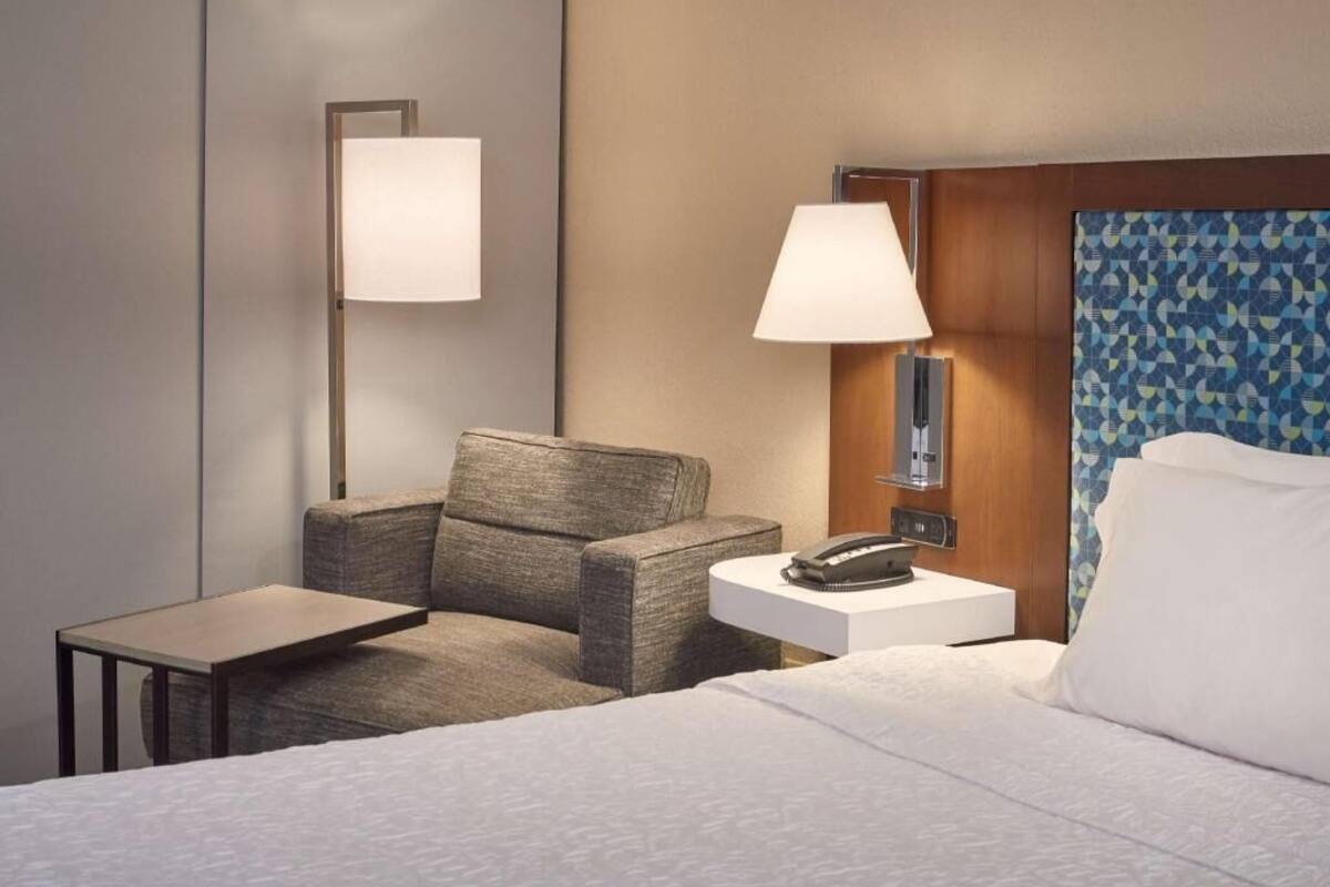 A hotel room with a bed, a chair, a small table, a bedside lamp, and a phone on a nightstand.