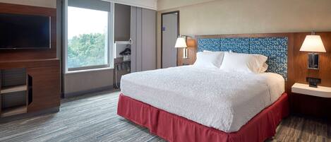 Habitación de hotel con una cama grande, un televisor de pantalla plana, una ventana con vistas a árboles y una cama con falda roja.