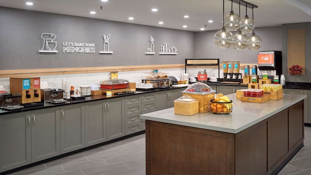 A modern kitchen with grey cabinets, a central island with fruit and snacks, and a wall with kitchen utensils and the phrase "Let's Bake Some Memories." - Hampton Inn by Hilton Harrisburg West (Mechanicsburg)