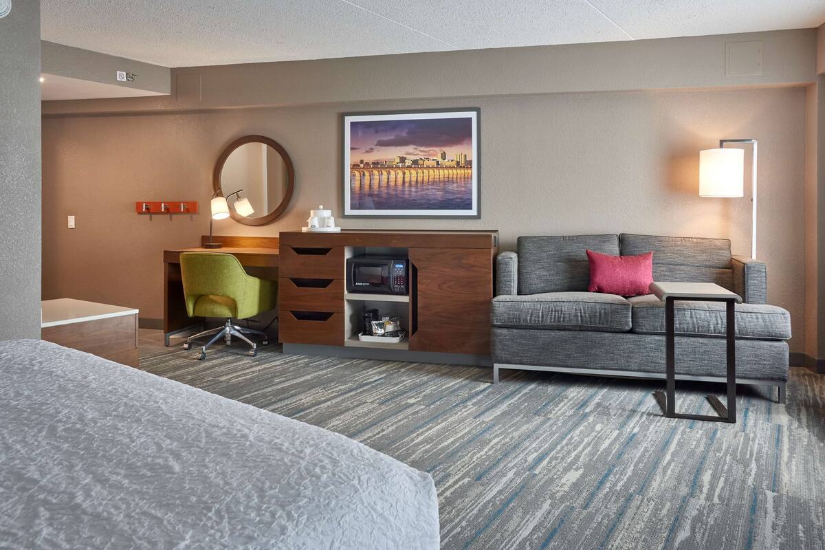 A hotel room with a bed, a desk with a chair, a grey sofa with a pink pillow, a framed picture on the wall, and a lamp.