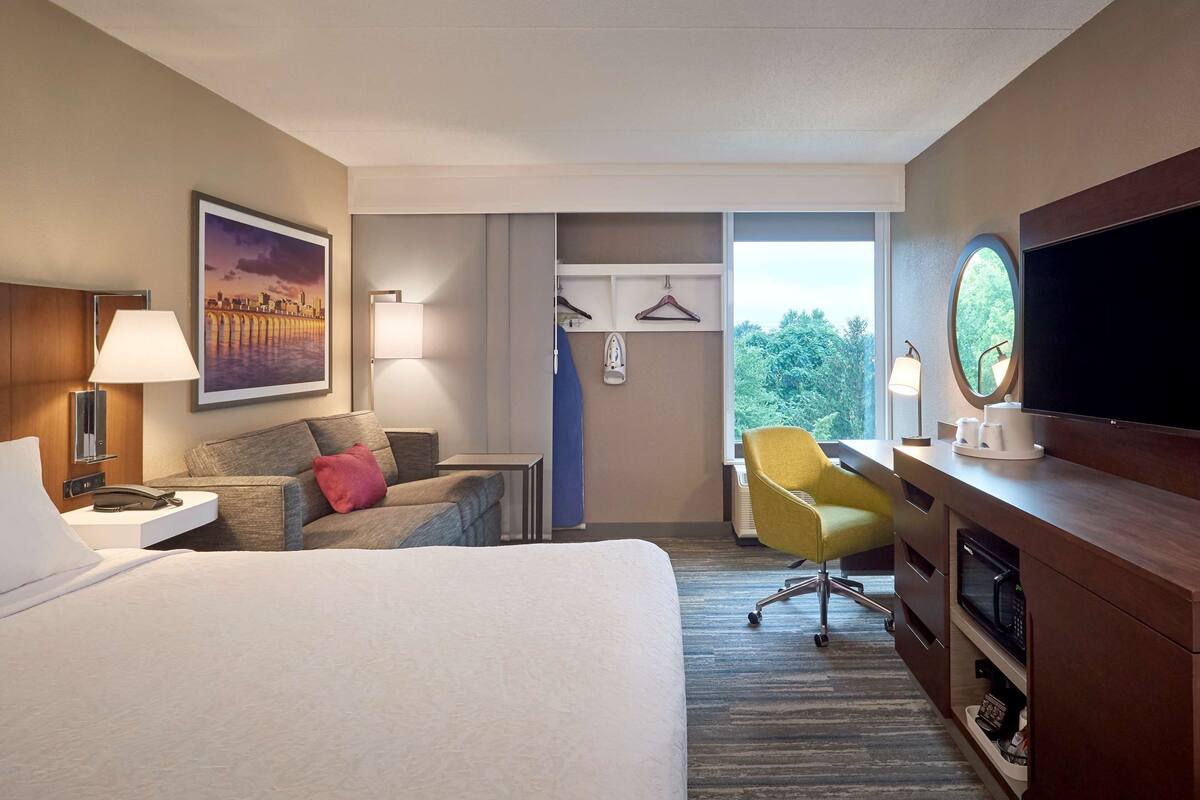 A hotel room with a bed, a desk with a TV, a chair, a desk lamp, a painting, and a view of trees.