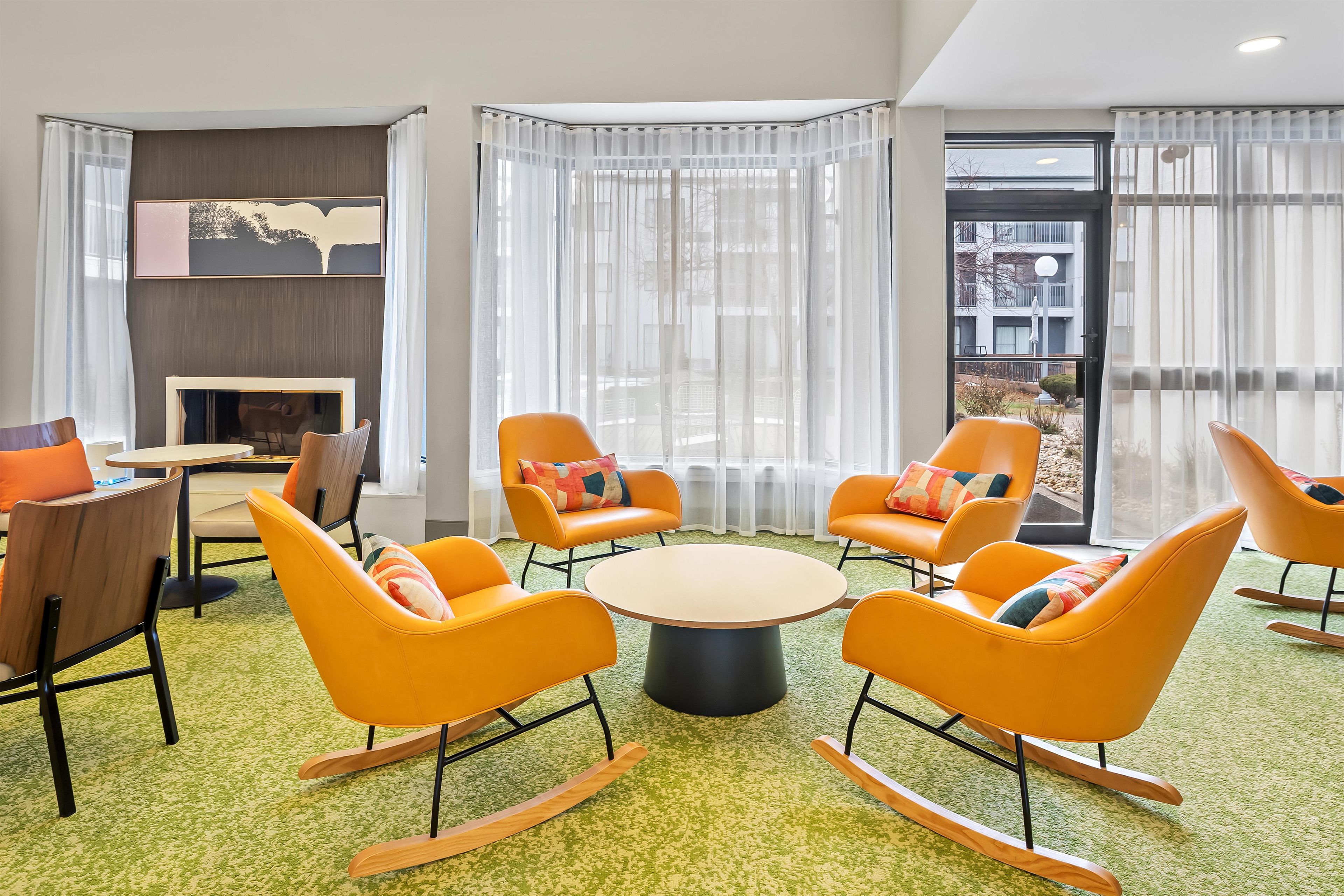 A modern lounge area with orange rocking chairs, a round table, and a fireplace.
