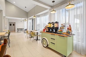 A modern kitchen area with a coffee station, dining tables, chairs, and a large window with sheer curtains.