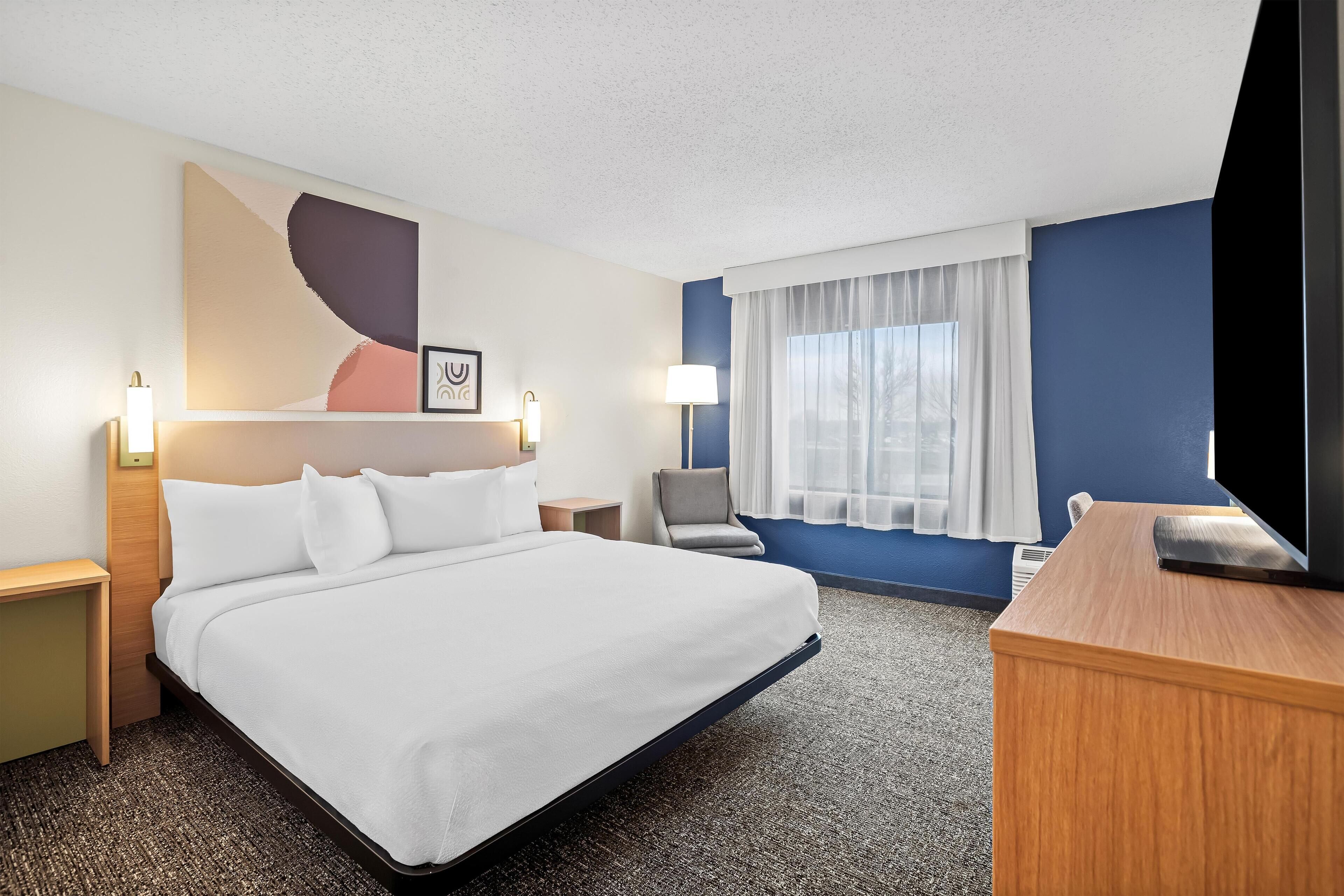 Une chambre d’hôtel avec un grand lit, un bureau, une chaise, une télévision et une fenêtre avec des rideaux.