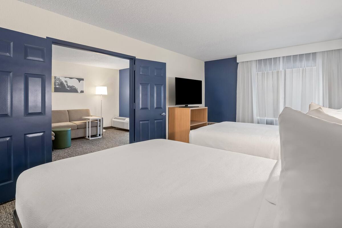 Une chambre d’hôtel avec deux lits, une télévision, un canapé, une lampe et une fenêtre avec des rideaux.