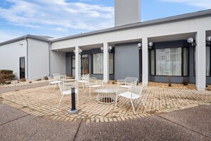 Un edificio moderno con un área de estar al aire libre techada, que presenta sillas y mesas de metal blanco.