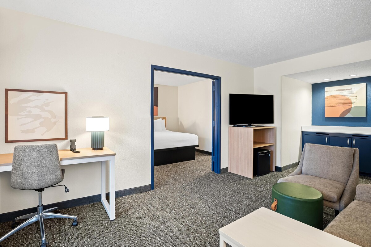 Une chambre d’hôtel avec un lit, un bureau, une chaise, une télévision et un petit pouf vert.