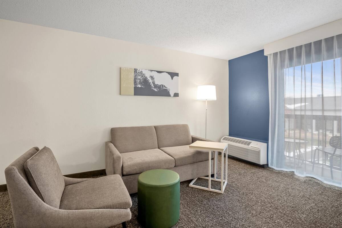Une chambre d’hôtel avec un canapé gris, un pouf vert, une petite table, une lampe sur pied et une grande fenêtre ornée de rideaux légers.