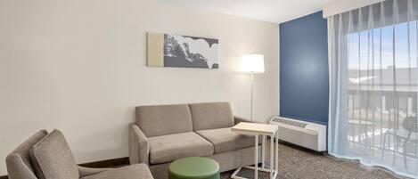 A hotel room with a grey sofa, a green ottoman, a small table, a floor lamp, and a large window with sheer curtains.