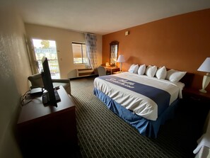 A hotel room with a bed, desk, and chair. There is a window with curtains, a lamp, and a mirror on the wall.