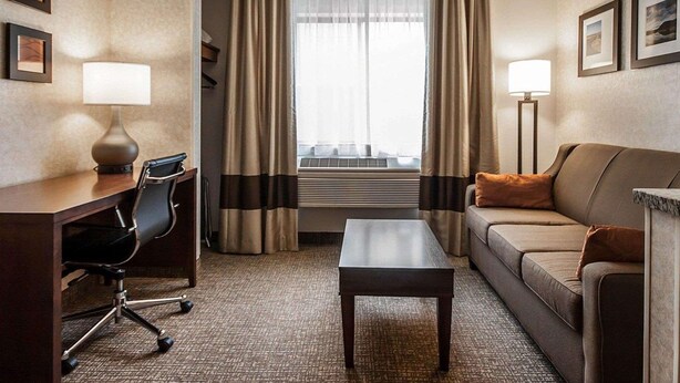 Una habitación de hotel con escritorio, silla de oficina, sofá y una mesita. Hay dos lámparas, una ventana con cortinas y una luz en el techo.