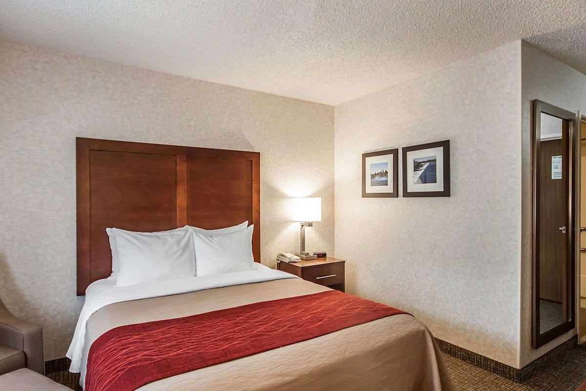 A hotel room with a wooden headboard, a bed with white and beige bedding, a red throw, a nightstand with a lamp, and two framed pictures on the wall.