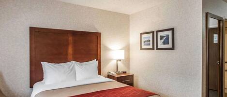 A hotel room with a wooden headboard, a bed with white and beige bedding, a red throw, a nightstand with a lamp, and two framed pictures on the wall.