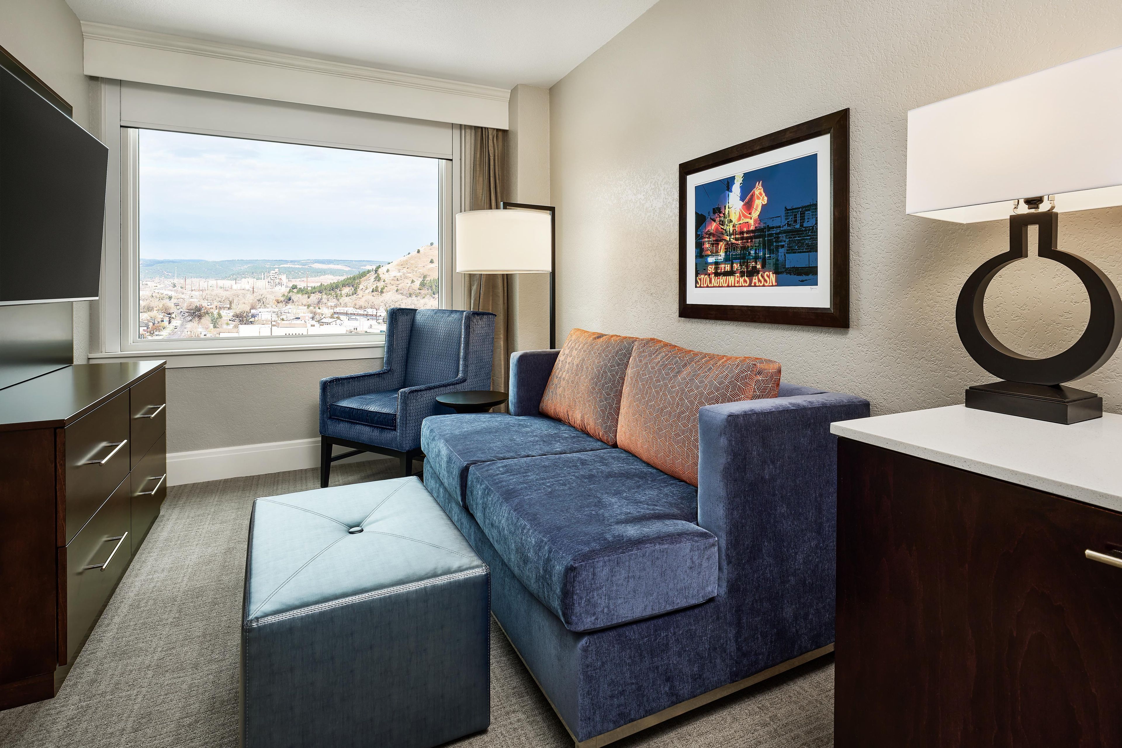 Quarto de hotel com uma janela grande, um sofá, uma poltrona, uma televisão, um abajur e um quadro na parede.