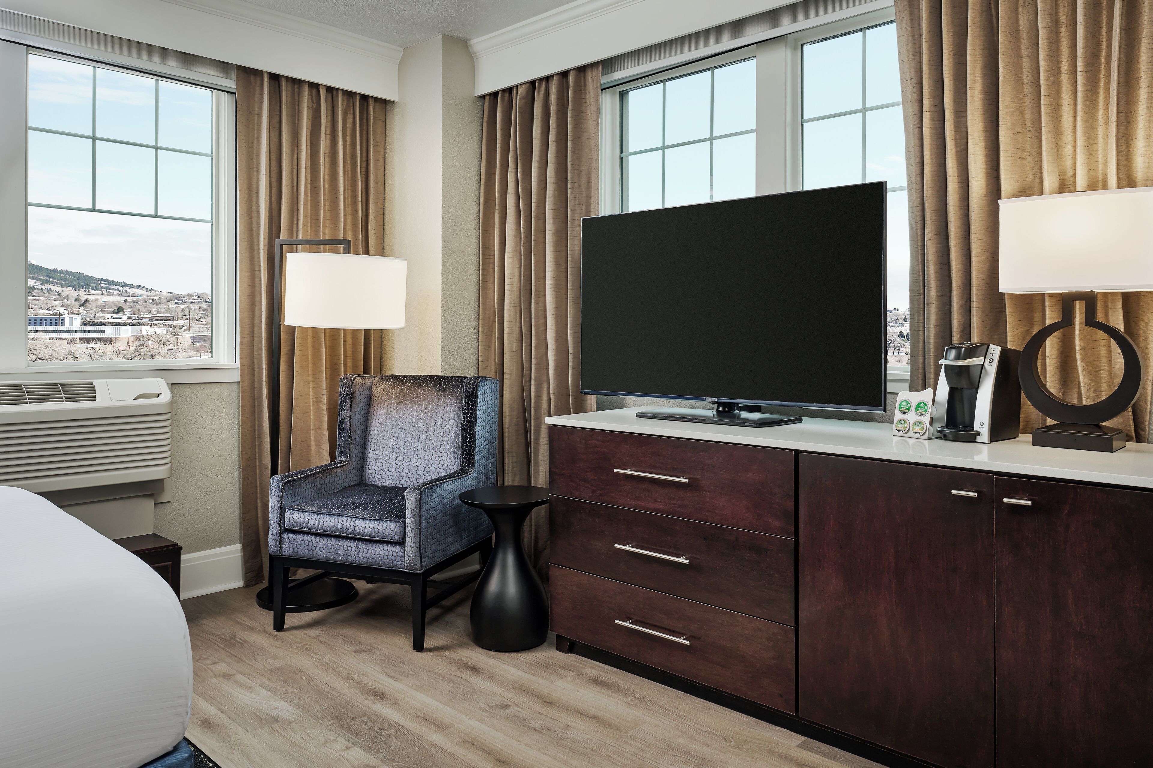 Une chambre d’hôtel équipée d’un grand téléviseur à écran plat, d’une chaise, d’une petite table d’appoint, d’une commode avec des tiroirs et d’une fenêtre avec des rideaux.