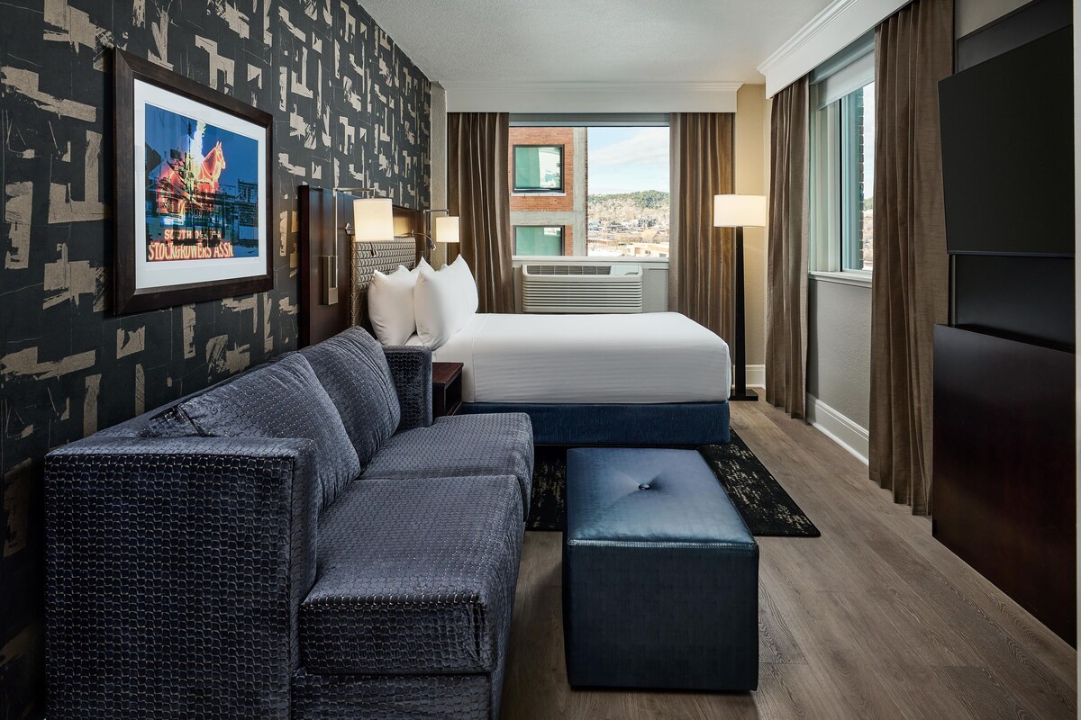 Quarto de hotel com uma cama grande, um sofá, um pequeno banquinho, uma escrivaninha com luminária e um quadro na parede.