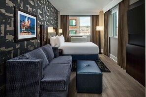 Une chambre d’hôtel avec un grand lit, un canapé, un petit pouf, un bureau avec une lampe et un tableau encadré au mur.