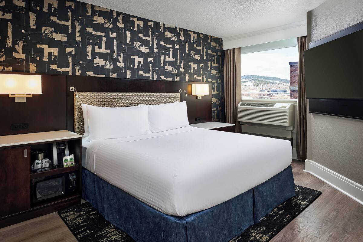 A hotel room with a large bed, a nightstand with a lamp, a built-in TV, a window with a city view, and a wall with a geometric pattern.