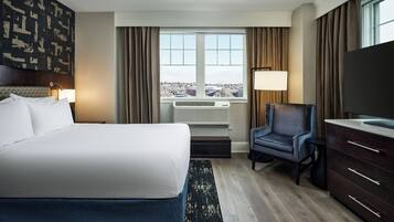 A hotel room with a large bed, a desk with a flat-screen TV, a chair, a dresser, and a window with a city view.