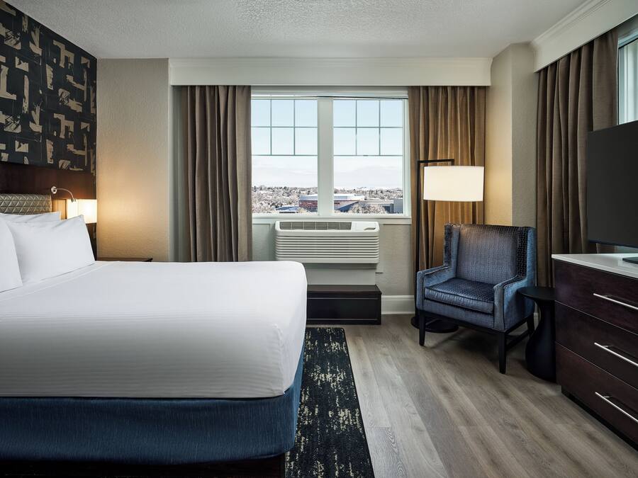 A hotel room with a large bed, a desk with a flat-screen TV, a chair, a dresser, and a window with a city view.