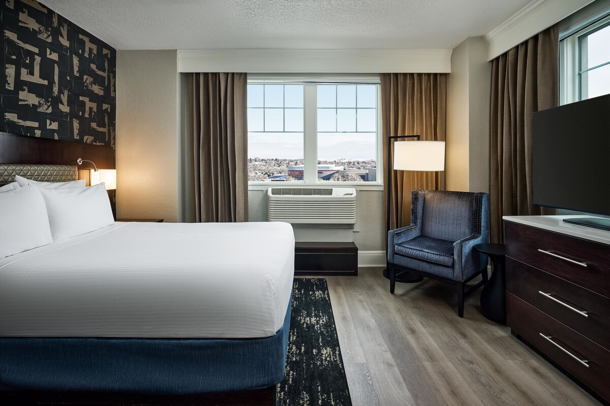 A hotel room with a bed, a desk, a chair, a TV, a window with a view, and a wall with a geometric pattern.